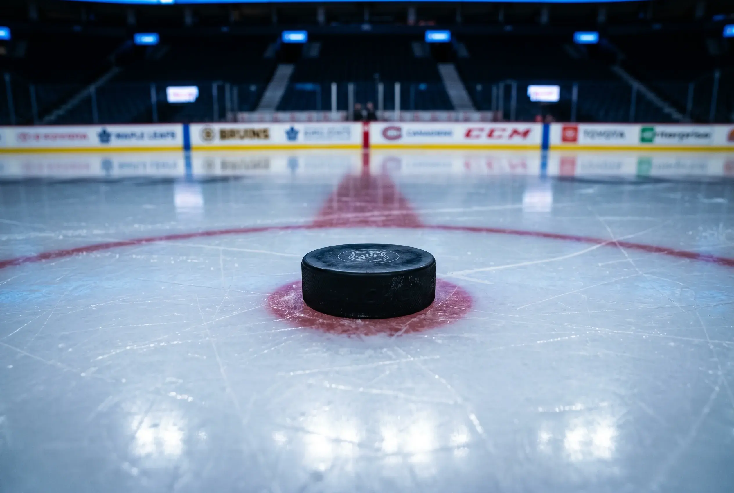 Naerbillede af en ishockey-puck pa en isbane med holdlogoer pa tavlen i baggrunden